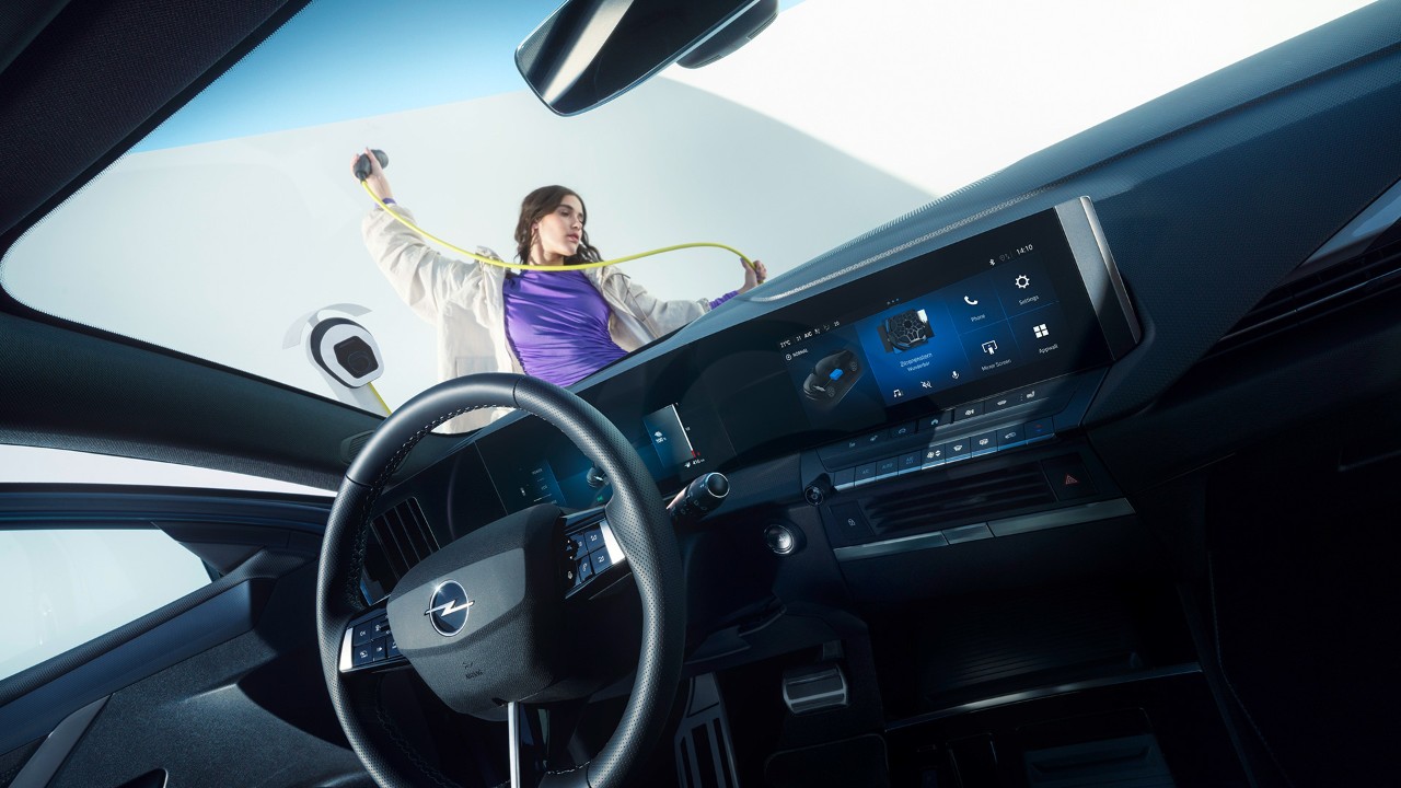 Interior view of an Opel Astra Electric from the passenger seat with woman holiding a charging cable outside