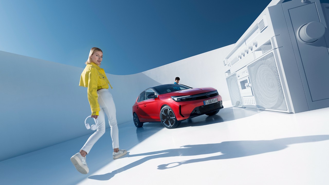 Femme debout à côté de la nouvelle Opel Corsa de couleur rouge avec toit noir et un homme qui monte par la porte du conducteur.