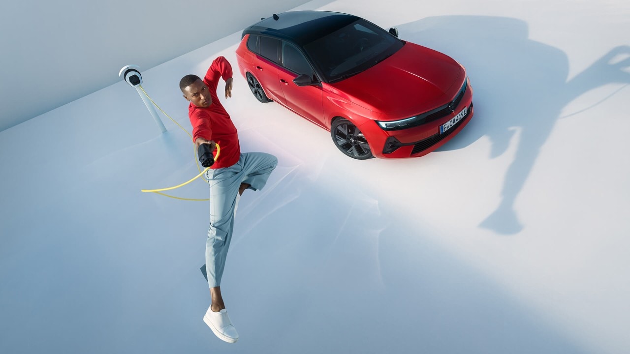 Man springt in de lucht met een EV-laadstation en een Opel Astra Sports Tourer Electric op de achtergrond