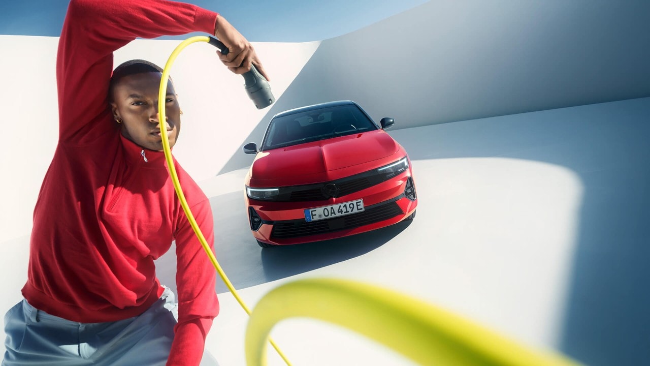 Man met oplaadkabel voor elektrische auto naast rode Opel Astra Sports Tourer Electric met zwart dak.