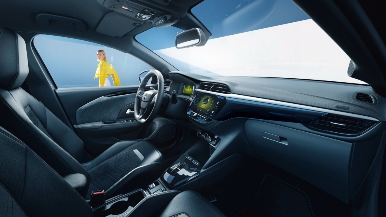 Opel Corsa Electric zwart interieur zijaanzicht vanuit passagierszetel met vrouw in gele t-shirt in de achtergrond. 