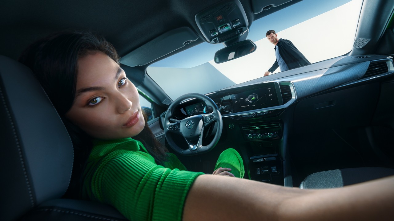Vrouw kijkt achterom terwijl ze op de bestuurdersplaats van een Opel Mokka Electric zit naast een man die naar buiten loopt