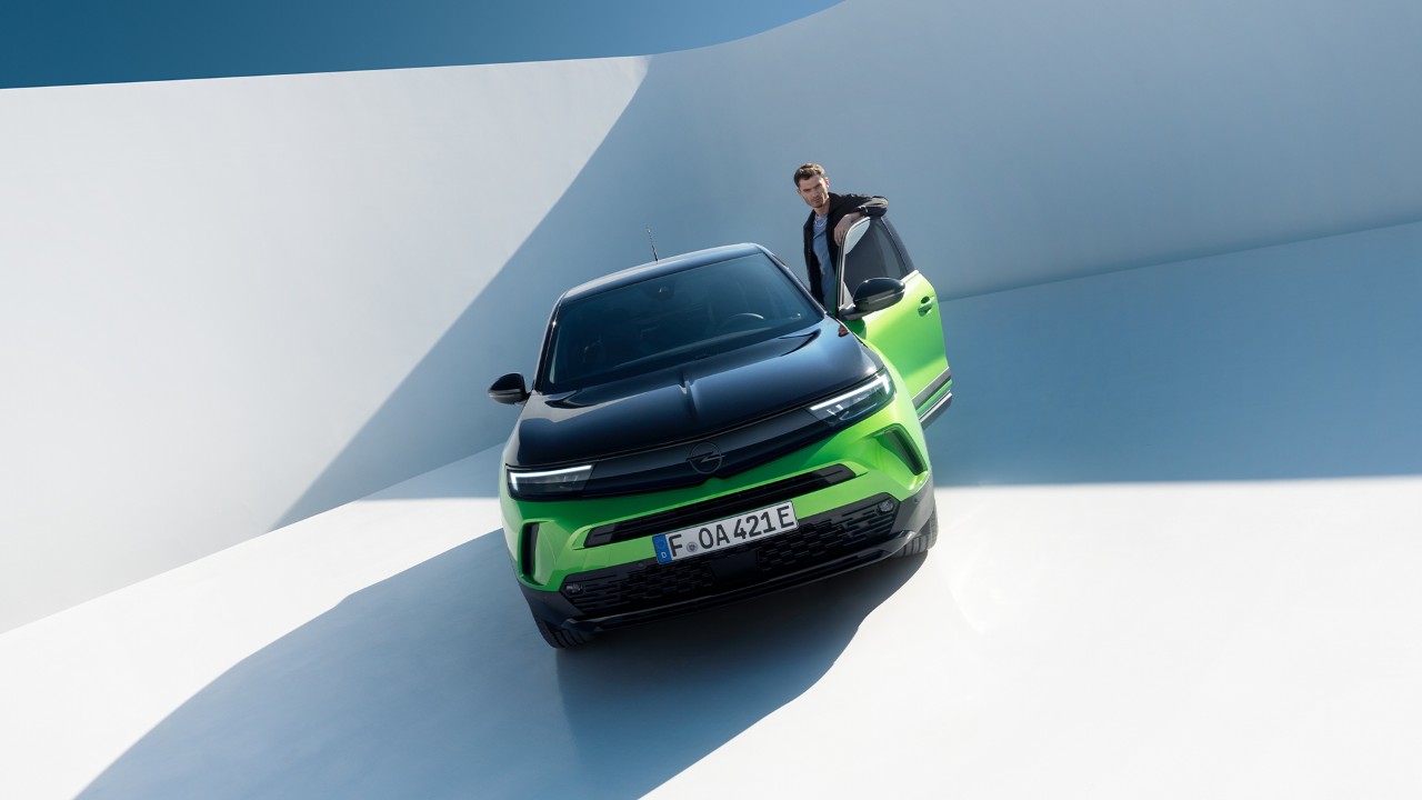 Vooraanzicht van een groene Opel Mokka Electric met zwarte velgen en een zwart dak met een man die instapt via de deur van de bestuurder.